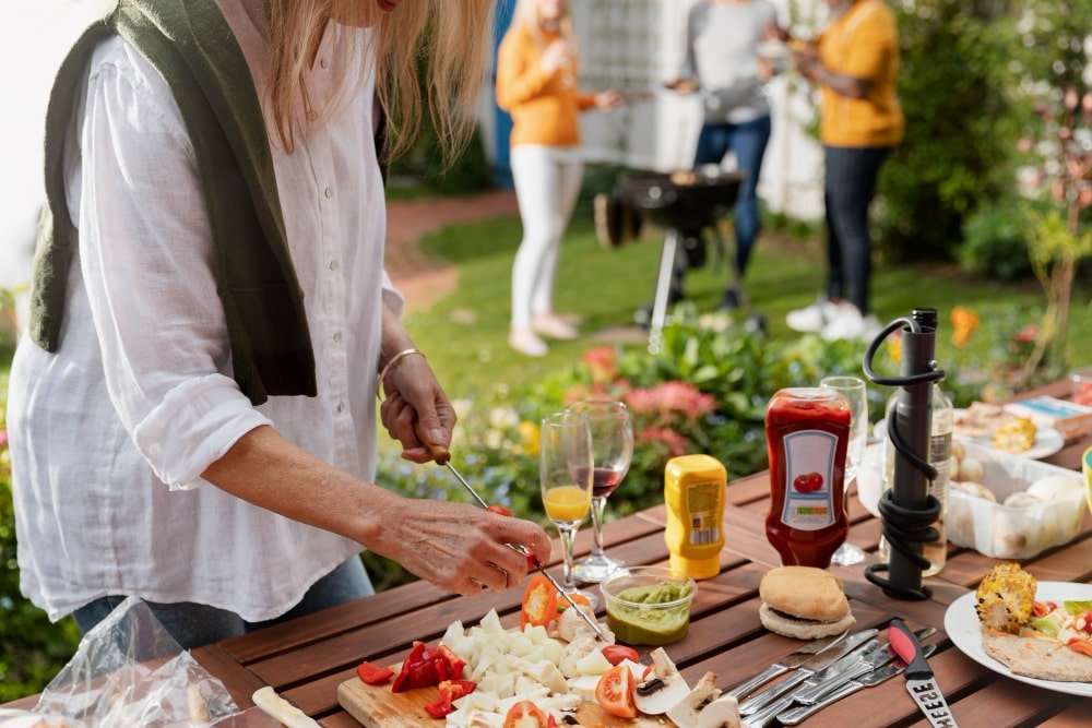 side-view-woman-making-skewers-min A woman preparing fresh skewers on a wooden table surrounded by condiments and drinks, while a group of friends gathers near a grill in a vibrant backyard setting in Etowah, NC, showcasing the functionality and charm of outdoor kitchens for entertaining.