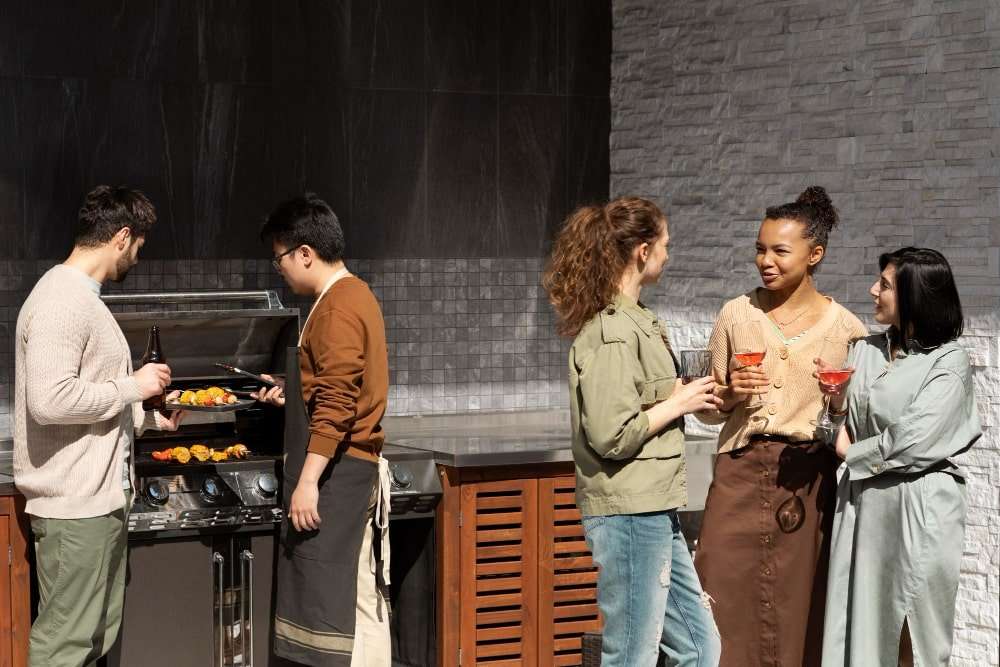 medium-shot-friends-making-barbecue-min A group of friends enjoying food and drinks around a stylish outdoor kitchen in Etowah, NC, featuring a built-in grill, custom countertops, and modern stonework, showcasing how outdoor kitchens enhance backyard spaces for entertaining.